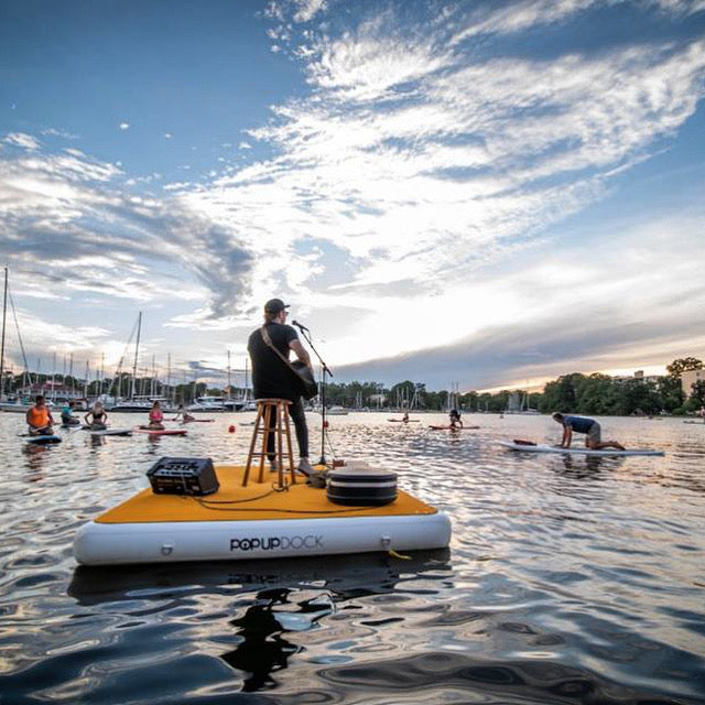 Man sings with guitar and microphone sitting on a stool on top of a floating PopBoard Inflatable Dock Platform to a crowd of paddleboarders onlooking the performance in the water.