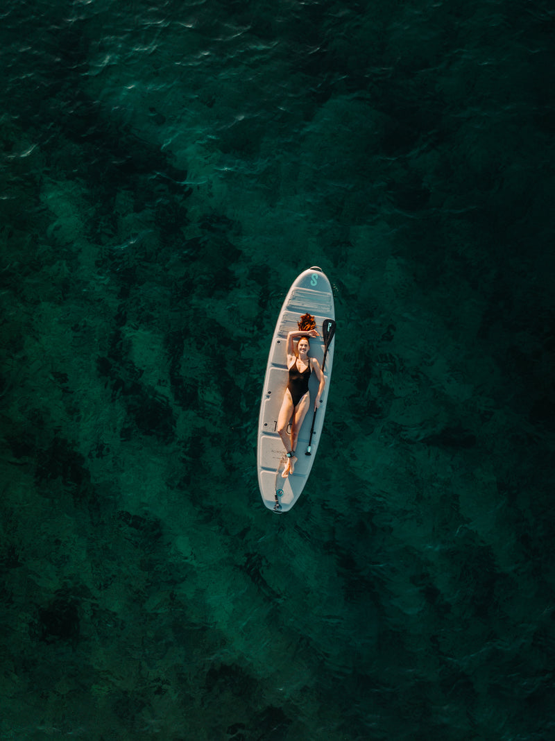 Woman laying down floating on SIPA Boards Alpha One Electric Paddleboard with self-inflating design and assist power unit floating on the water with sailboats. Ships free from e-boarders.com.