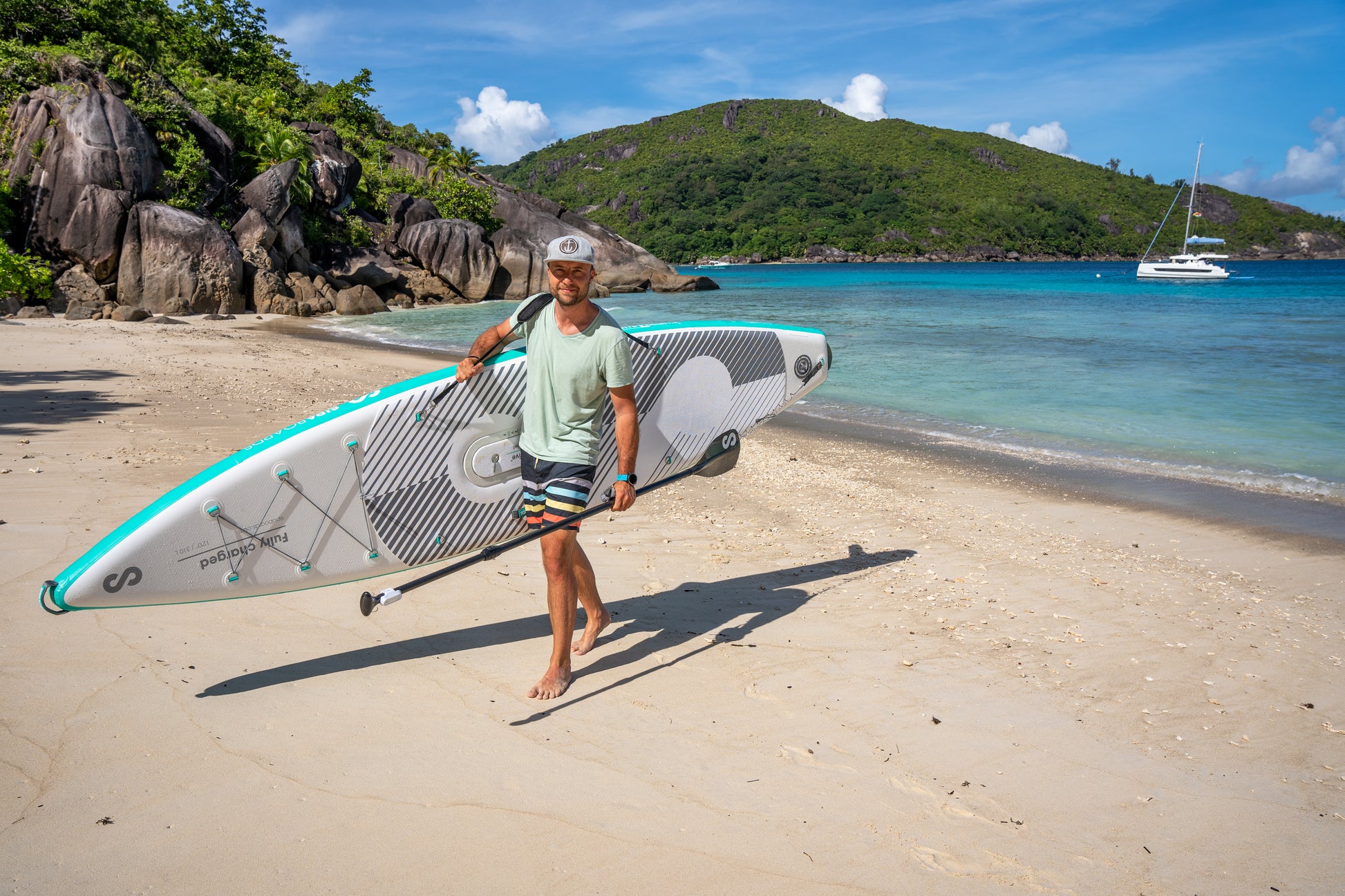 Man on beach carrying SIPA Boards Alpha One Electric Paddleboard with self-inflating design and assist power unit floating on the water with sailboats. Ships free from e-boarders.com.