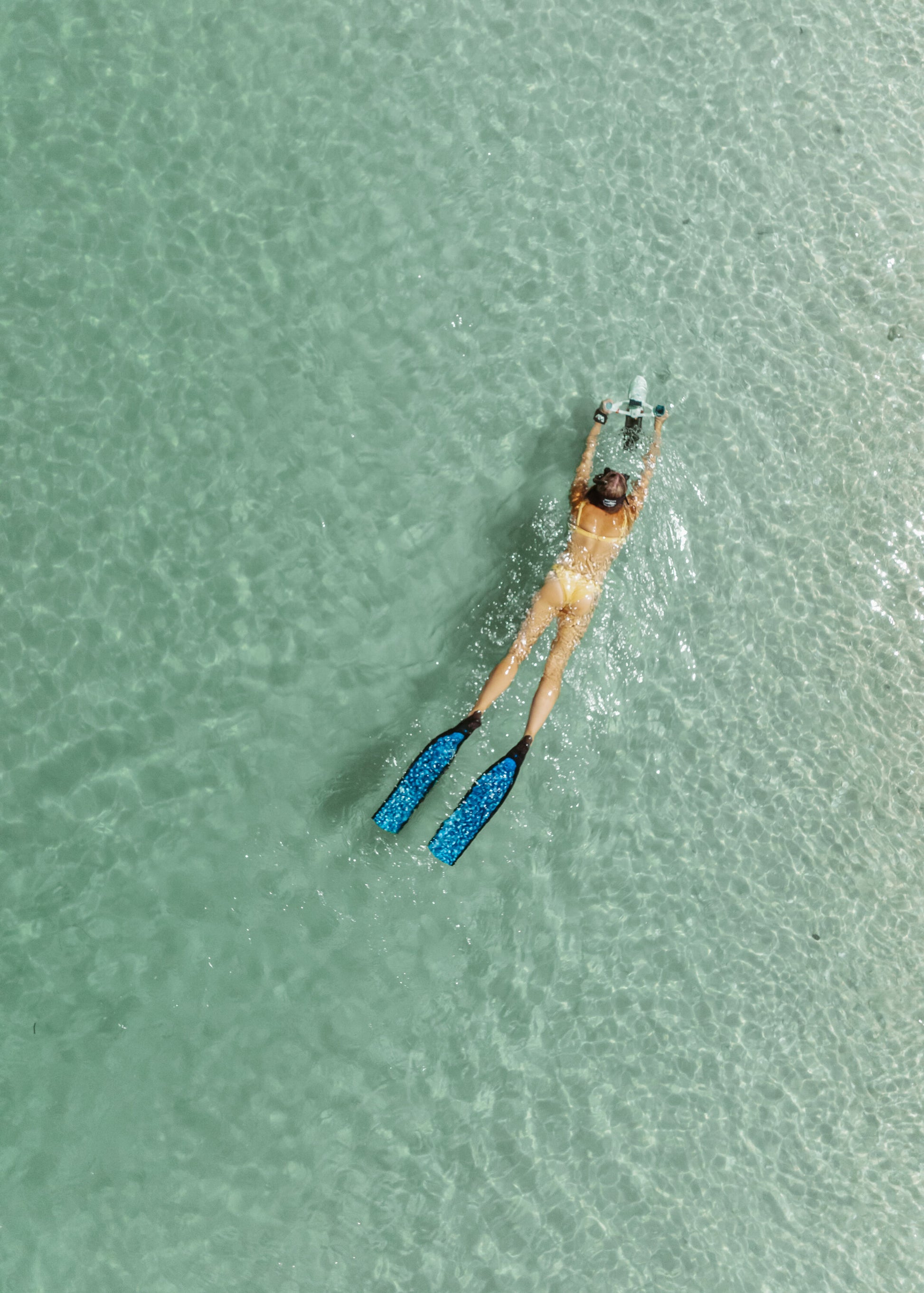 Woman swimming in shallow ocean waters with Aqua Marina BLUEDRIVE X Pro electric-assist fin system. Includes wireless remote control and rechargeable battery. Designed for paddleboarders, snorkelers, and recreational swimming. Free shipping at e-boarders.com