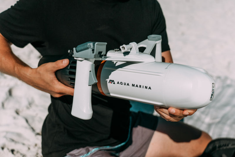 Man holding Aqua Marina BLUEDRIVE X Pro electric-assist fin system. Includes wireless remote control and rechargeable battery. Designed for paddleboarders, snorkelers, and recreational swimming. Free shipping at e-boarders.com