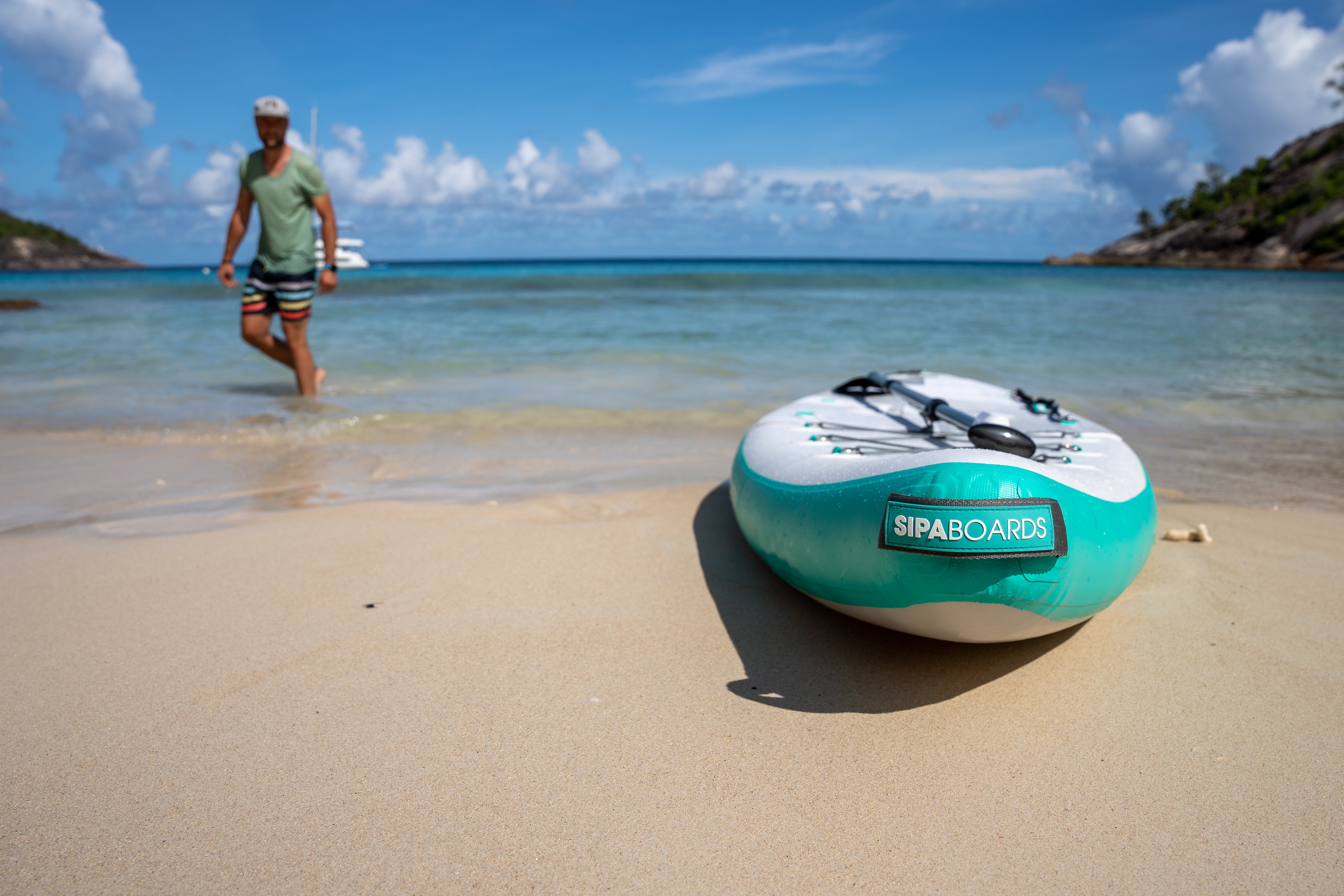 Man on beach with SIPA Boards Alpha One Electric Paddleboard with self-inflating design and assist power unit floating on the water with sailboats. Ships free from e-Boarders.com.